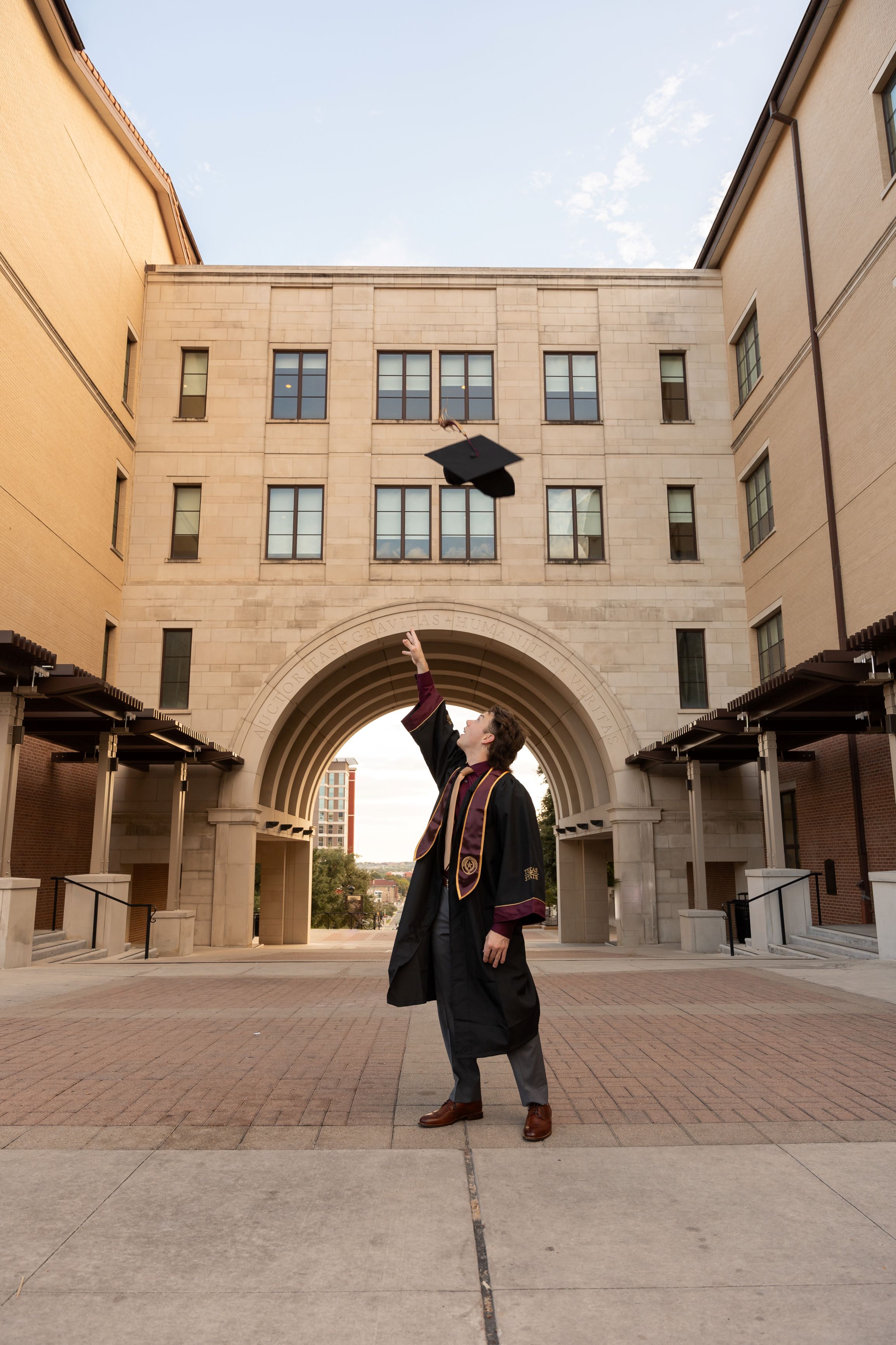 Jack in his graduation gown, throwing his cap in the air in front of Texas States UAC Building known as 'the arch'